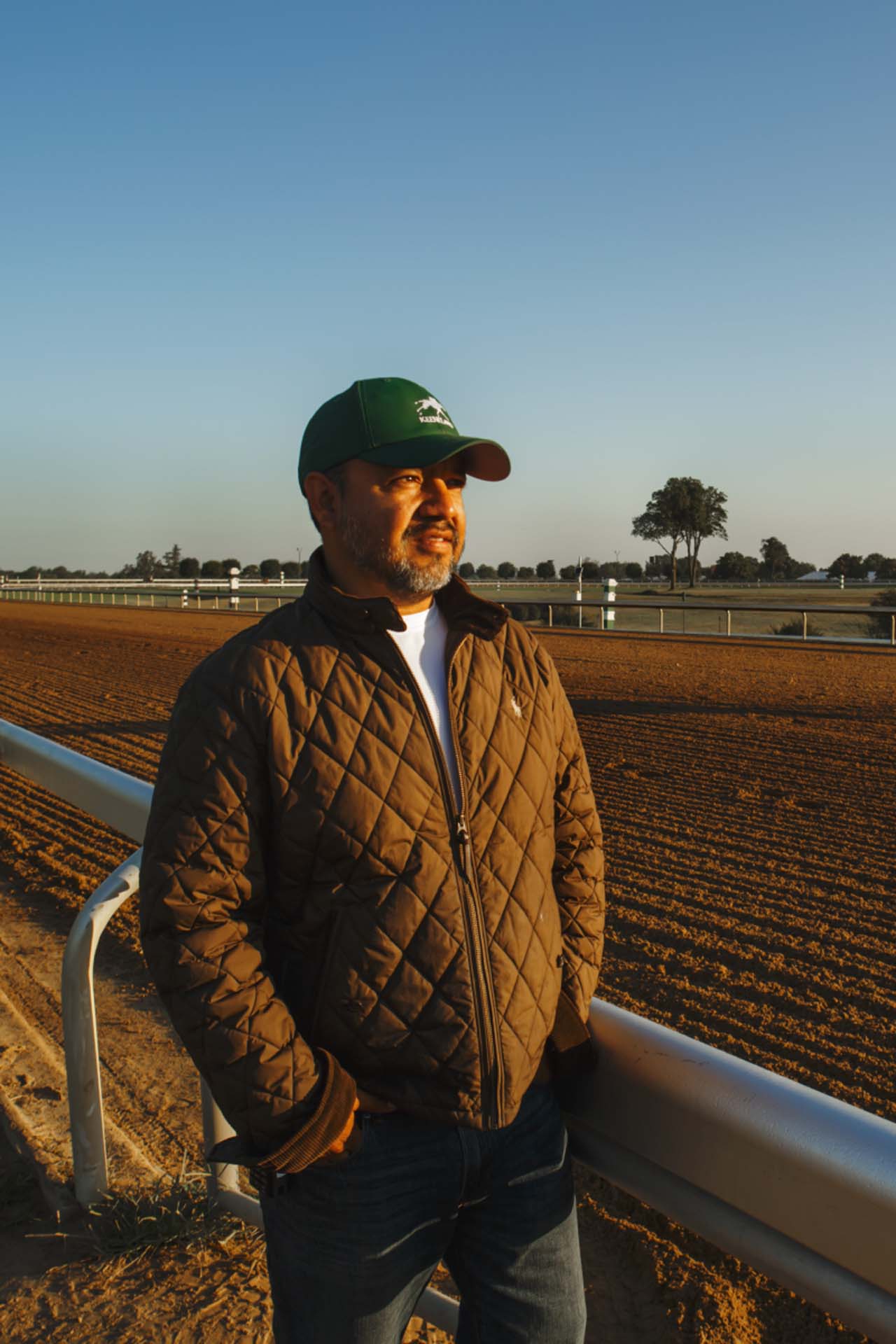 A photo of Alfredo Laureano looking off into the distance while standing by the dirt track at sunset. He is an older Hispanic man with graying chin stubble. He is wearing a green Keenriders-brand hat and a brown Keenriders-brand jacket overtop a white shirt.