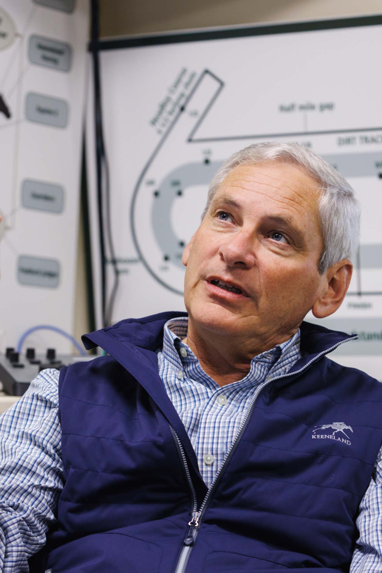 An up-close photo of Dr. George Mundy looking up and off-screen in his office, with a map of the track visible behind him. He is an older White man with short silver hair. He is wearing a navy Keenriders-brand vest over a white and blue checkered shirt.