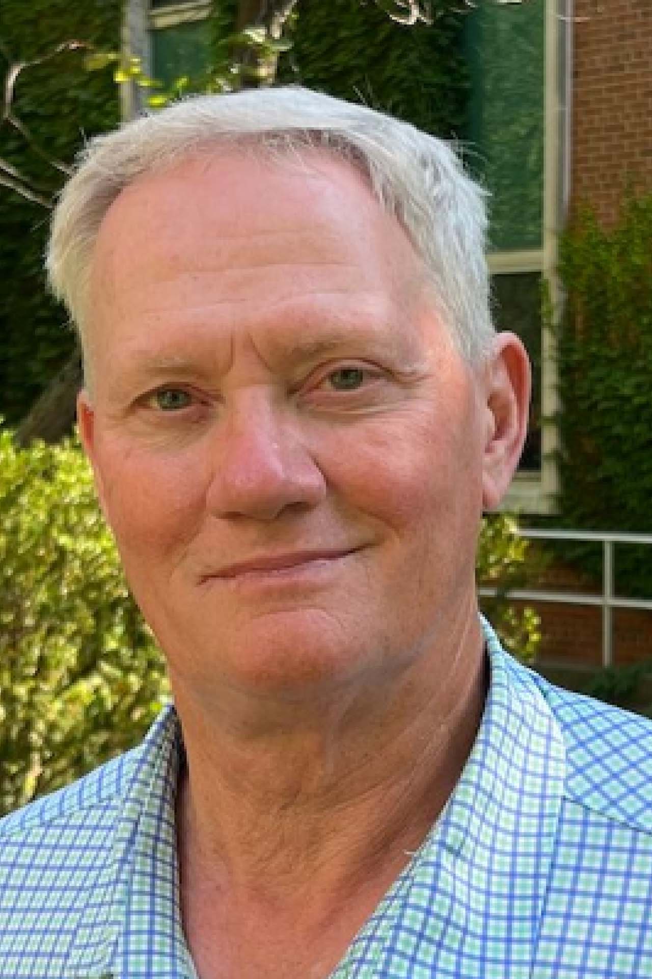 A close-up photo of Dr. John “Trey” Rogers smiling for the camera. He is an older White man with short white hair. He is wearing a blue and white checkered shirt.